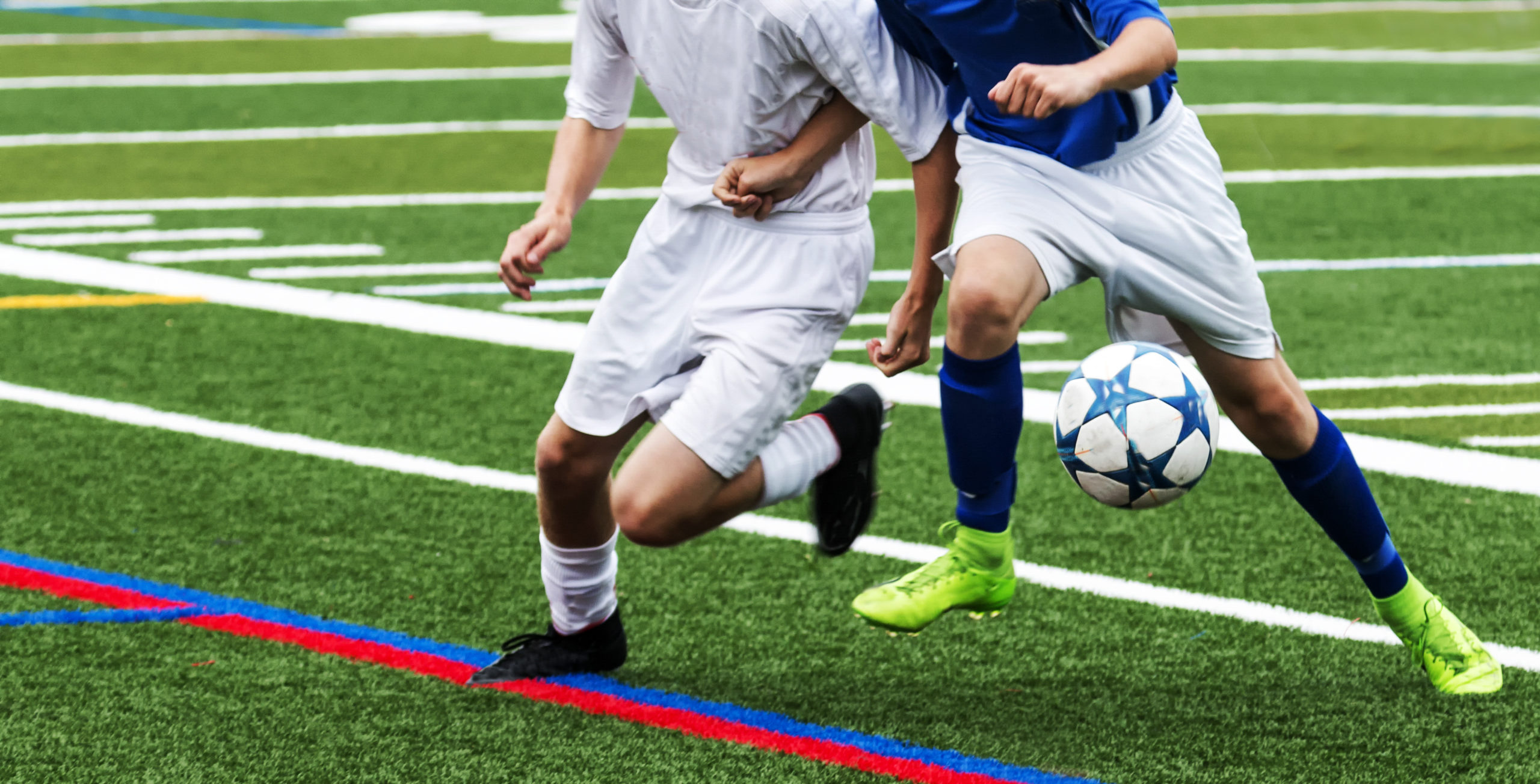 Two high school boys soccer players pushing and fighting each other to get controll of the soccer ball on a green turf field.